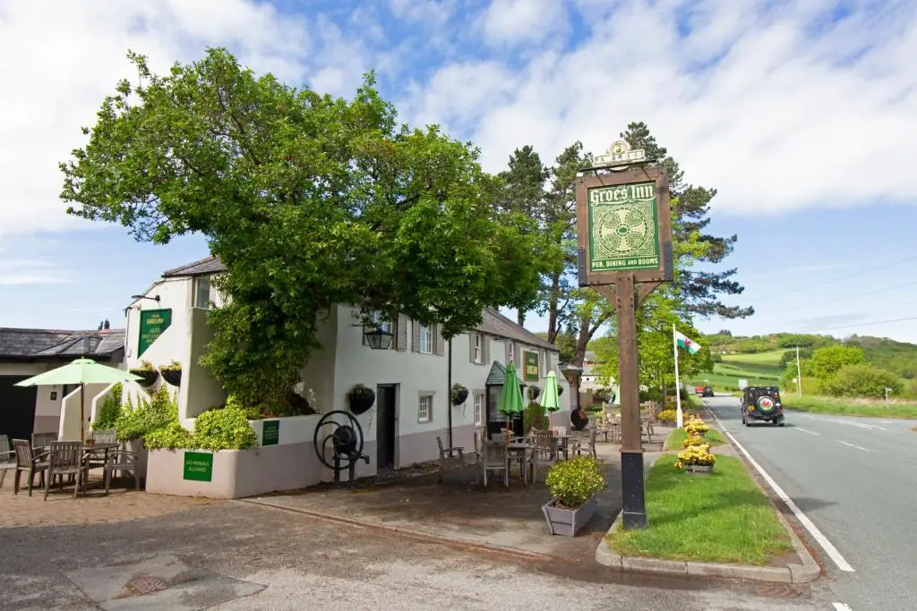 A short, wide, light-coloured building with some benches around the side and front. There's branches covering the roof, and a big standalone sign on a small patch of grass at the front stating "Groes Inn"