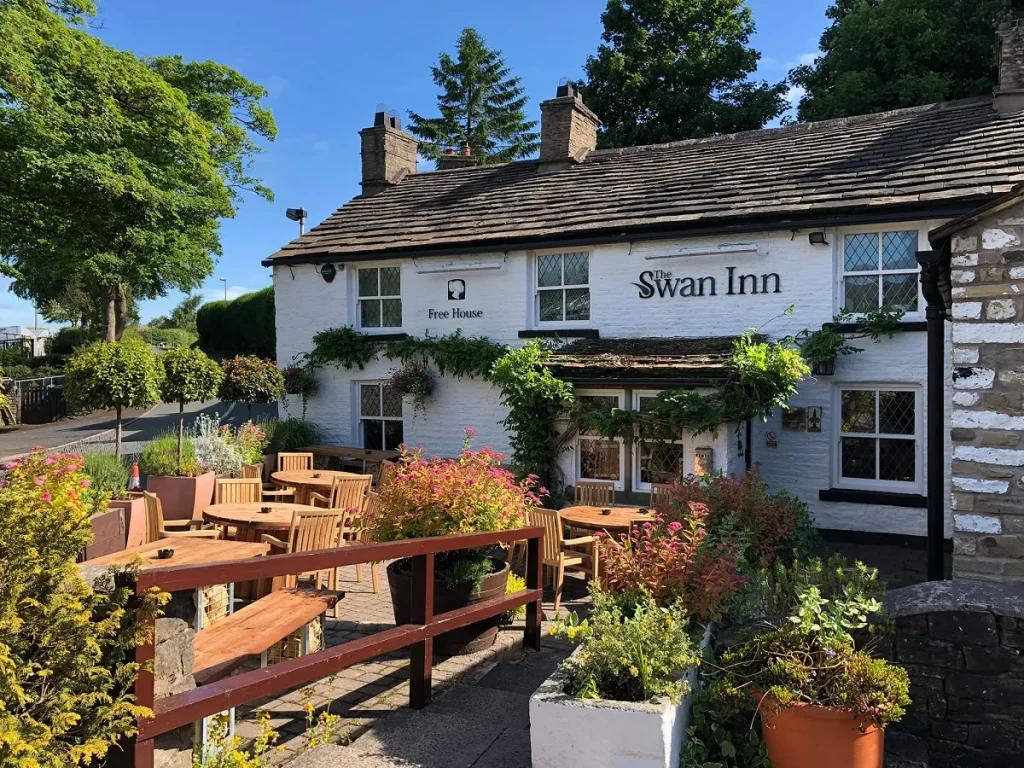 A picture of the The Swan Inn on a sunny day. Old tudor-style looking building with lots of bright flowers and tables outside.