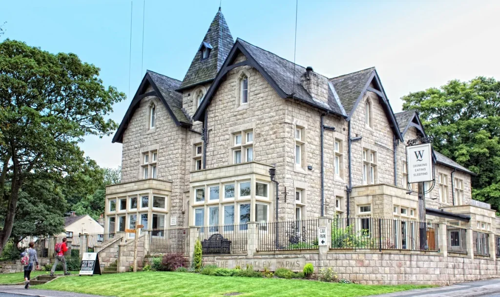 A large light coloured building with small patches of grass and some stairs to the side. There's a sign tucked behind the shrubbery that says "The Wheatley Arms"