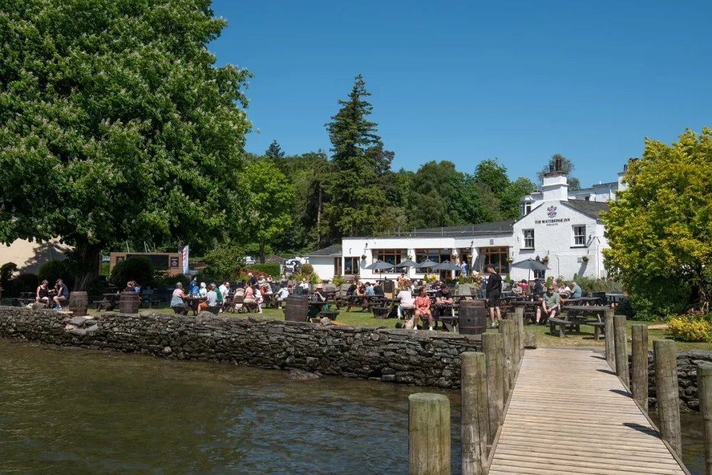 A picturesque image of the Wateredge Inn showing the river running alongside with plenty of guests enjoying the garden. Bright blue sky with lots of green trees.