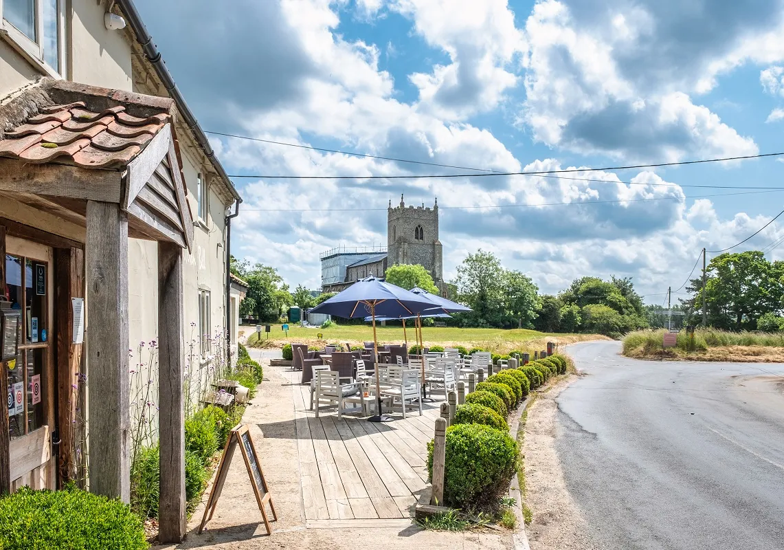 The front of the Wiveton Bell on a partly sunny/partly cloudy day, showing tables, parasols and a church in the background. Best Pubs with Rooms in Norfolk.