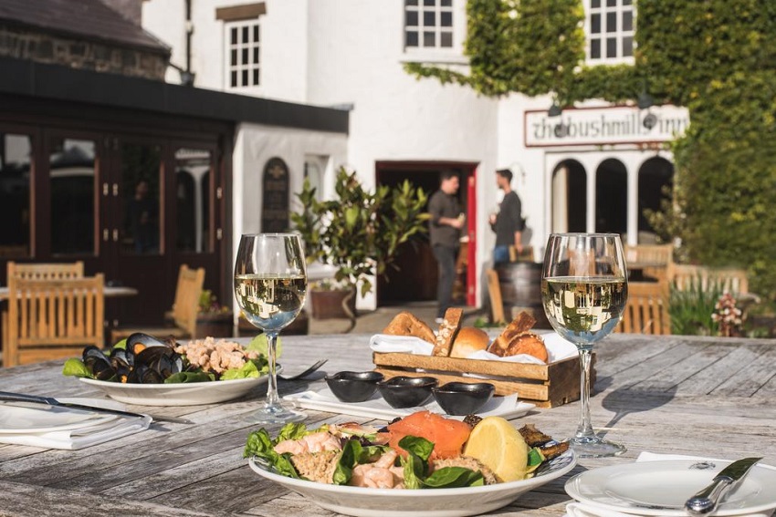 A close-up picture of 2 plates with food on and 2 wine glasses. The Bushmills Inn can be seen in the background, with lots of shrubbery around the sign