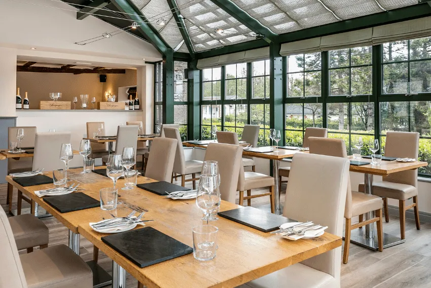 The Gamekeeper Inn's luxury restaurant - big, open glass windows, beige tables, cutlery and glasses on the tables. Bar area in the background.