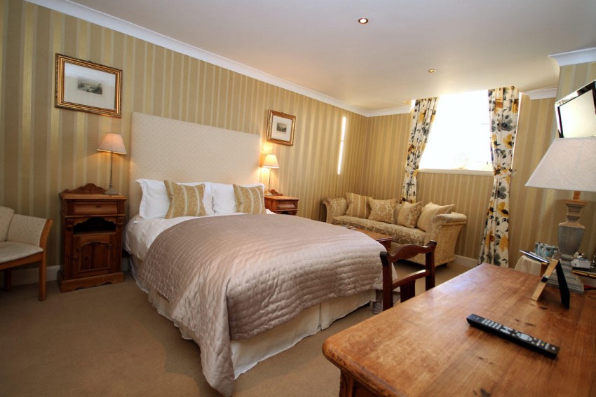 A luxury double bedroom with a sofa, bedside tables either side, lamps, desk with remote control and floral curtains that have been drawn to let the sunlight in